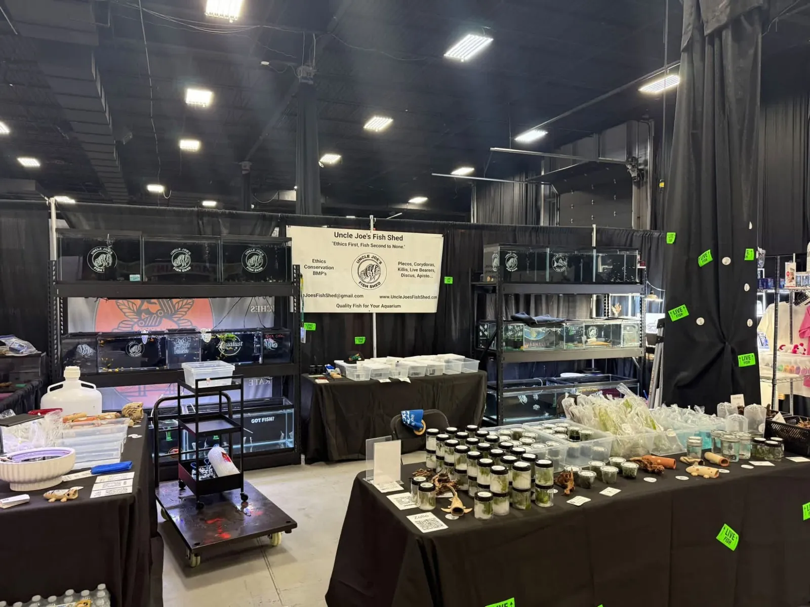 Booth at Aquashella 2026 in New Jersey with rows of fish tanks on black shelving, a banner for Uncle Joe's Fish Shed, and tables of aquatic plants in jars